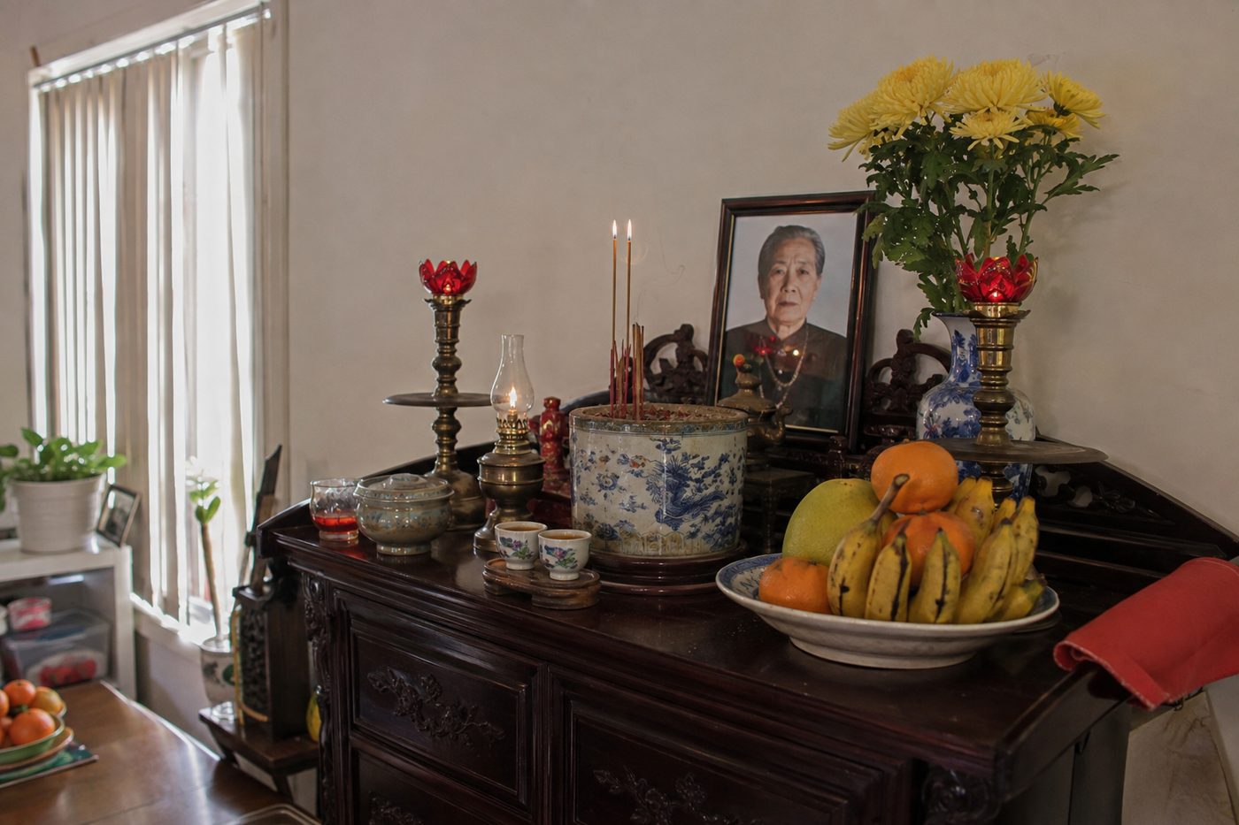 A Vietnamese family ancestor altar at home, with a framed photograph, burning incense, yellow chrysanthemums and offerings of fruit